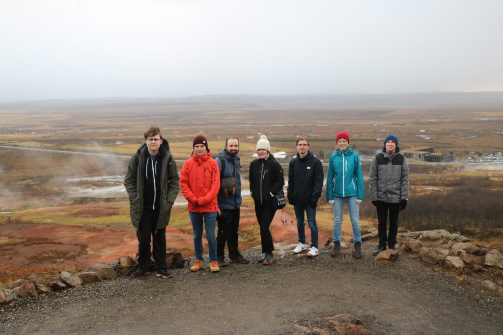Seitsemän ihmistä rivissä, taustalla islantilainen maisema, sää harmaa