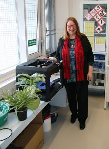 Maija Aksela laboratoriossa, takana kemikaalikaappi, edessä viherkasveja ja tekninen laite.