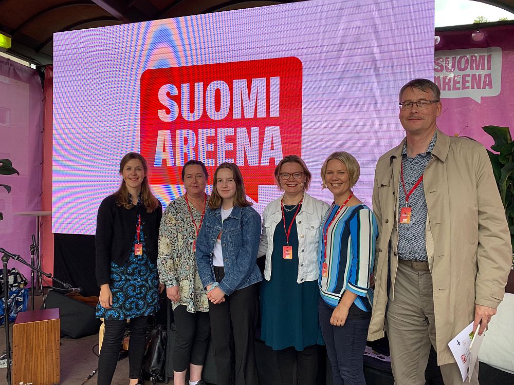 Kuusi henkilöä poseeraa Suomi Areena -näytön edessä