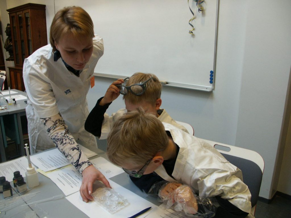 lapsia laboratoriotakeissa pöydän ääressä tutkimassa eri nesteitä sekä ohjaaja seisomassa pöydän vieressä