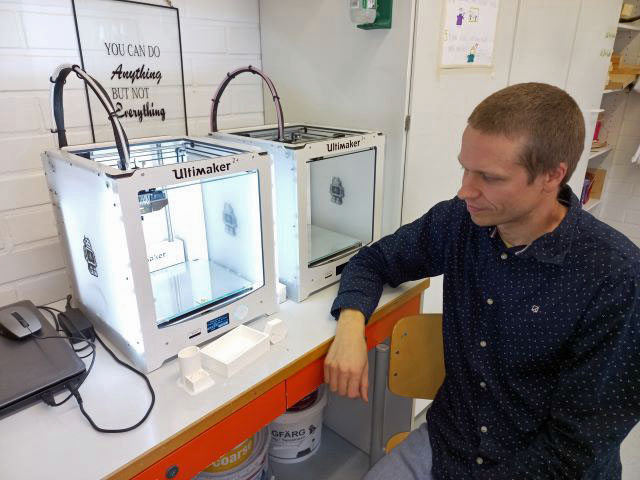 Henrik Vähäkangas noja apöytään ja katsoo kahta 3d-tulostinta jotka ova tpöydällä. Miten 3D-tulostusta voi hyödyntää eri oppiaineissa.