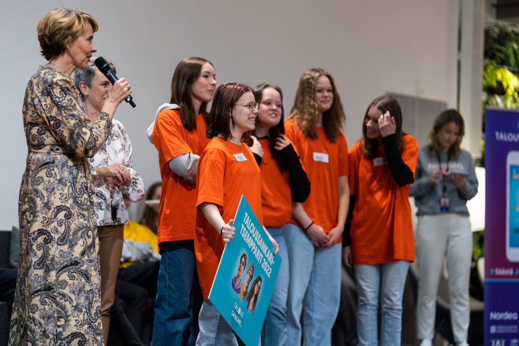 Palkintojenjakotilaisuus. Oranssipaitainen teinitytöistä koostuva joukkue, sekä muutamia muita henkilöitä, keski-ikäinen nainen puhuu mikrofoniin.