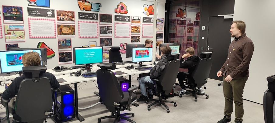 värikkäässä tietokoneluokassa neljä oppilasta tietokoneilla ja opettaja seuraa työskentelyä