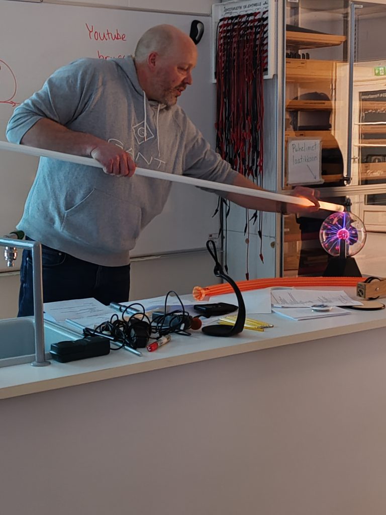 Mikko opettajanpöydän takana, käsissään loisteputkilamppu, jonka plasmapalloon koskeva pää hehkuu valoa. demoja ja kokeellisia töitä opetuksessa.